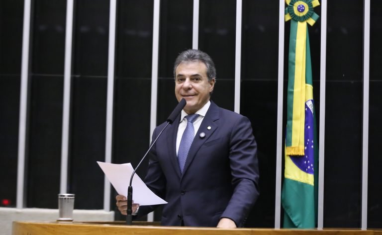 Deputado Beto Richa fala ao microfone. Ele é um homem claro, com cabelo escuro, usa um terno azul-marinho, camisa branca e gravata azul. Ele segura um papel. No fundo, à direita, há uma bandeira do Brasil